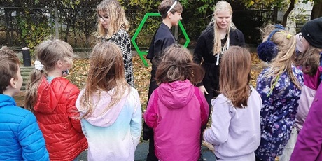 Powiększ grafikę: Na zdjęciu zajęcia w plenerze. Grupa dzieci słucha wolontariuszki która objaśnia zasady gry.